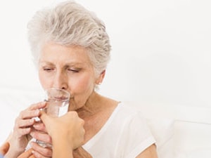 Clinician assists an older adult woman with dysphagia in drinking water during a bedside swallow assessment.