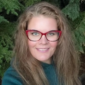 A smiling woman with long, wavy hair and red glasses stands in front of green pine trees.