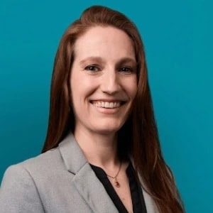 A woman with long brown hair in a gray blazer smiles in front of a teal background, representing medbridge.