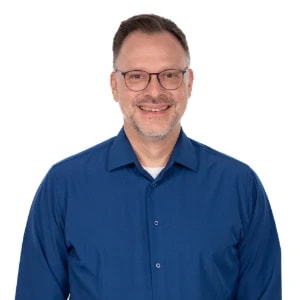 A man with glasses and a trimmed beard in a blue shirt smiles against a white background, representing the future of healthcare education with medbridge.