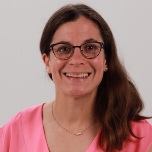 A woman with glasses and long brown hair smiles in a pink top, wearing a small necklace against a light background.