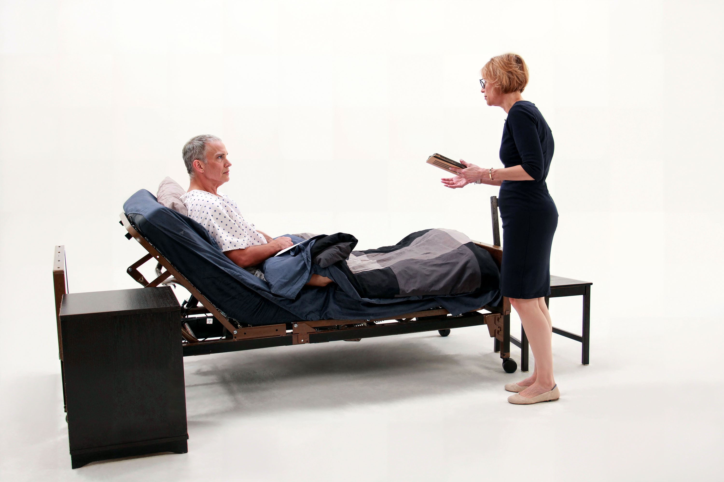 A man in a hospital bed talks to a woman with a clipboard, set against a plain background.