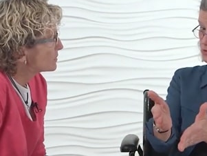 Two people conversing, one gesturing, against a textured white wall backdrop.