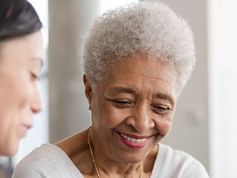 Two women, one younger and one older with short gray hair, chat and smile indoors in a casual or professional setting.