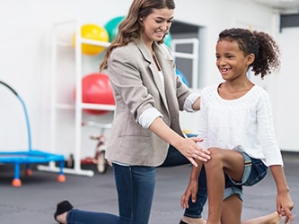 A therapist helps a girl with a knee exercise amid colorful therapy equipment.