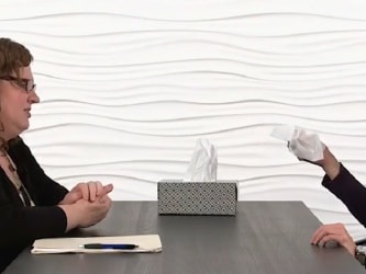Two women conversing at a table with tissues, papers, and a pen; medbridge-style wall backdrop.