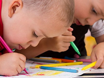 Two children color with markers at a table filled with art supplies.