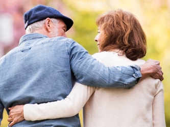 An elderly couple strolls through a vibrant park with pink blossoms and greenery.