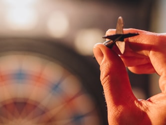 A hand ready to throw a dart with a blurred dartboard behind.