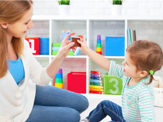 A woman and child play with toys on the floor in front of a colorful shelf displaying numbers.