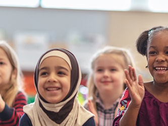 Children in a classroom smiling and clapping with blurred elements in the background.