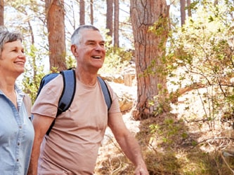 An older couple walks on a sunlit forest trail, with the man carrying a backpack.