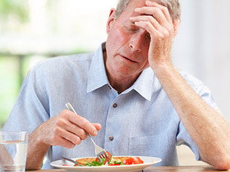 An elderly man in a light blue shirt sits at a table looking tired or stressed with food and water nearby.