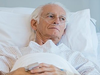 Elderly man in a hospital bed with an oxygen tube, wearing a gown, holding a device.