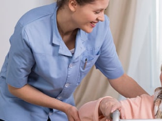 A Medbridge caregiver in blue helps a smiling elderly woman with a walker in a bright room.
