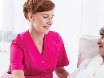 A nurse in pink chats with an elderly patient in bed at Medbridge.