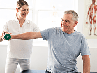 A woman helps an older man with a dumbbell exercise in a well-lit room at medbridge.