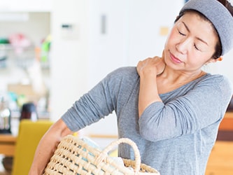 A woman in a gray shirt, holding a laundry basket and rubbing her neck, stands among wooden furniture.