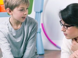 A boy and woman sit talking on the floor in a bright room with colorful furniture and toys.