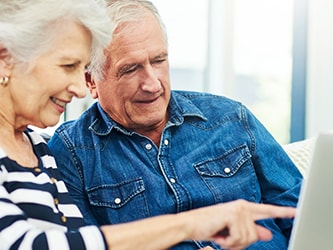 An older couple uses a laptop together in a bright room, with the woman pointing at the screen.