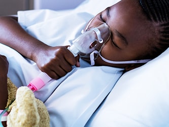 A child in bed with a nebulizer mask and holding a teddy bear attached to an IV drip.