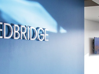 Office interior with a blue wall featuring the medBridge logo, a window, and a wall-mounted TV on the right.