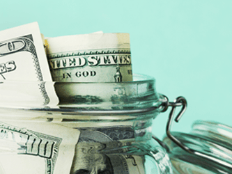 A glass jar with U.S. dollar bills is open on a light turquoise background.