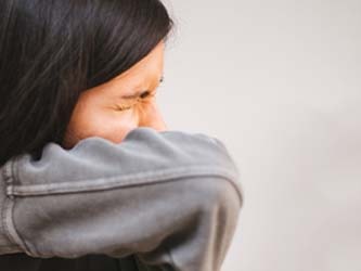 Person in a gray sweatshirt sneezing into their elbow with a plain background.