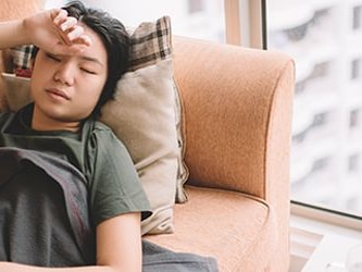 Someone resting on a sofa with a blanket by a window, hand on forehead.