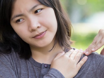 Individual in a long-sleeve shirt outdoors, touching their shoulder with a thoughtful look.