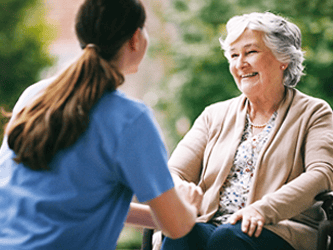 A caregiver in blue scrubs smiles beside an elderly woman in a wheelchair outdoors.