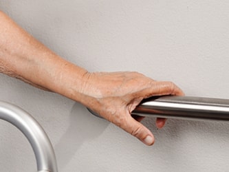 An elderly hand holds a wall-mounted metal handrail beside a walker.