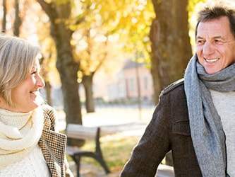 A man and woman in scarves and coats smile at each other while walking on a path lined with autumn leaves.