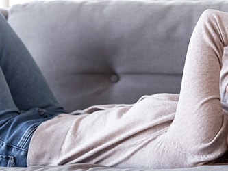 A woman in a beige sweater and blue jeans is lying on a grey couch with her hands on her head.