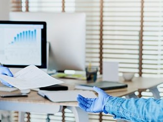 Masked and gloved individuals exchange a document in an office with computers and large windows.