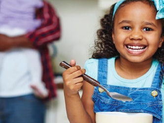 Child in overalls at kitchen counter smiles with bowl and spoon, while adult with baby is in background.