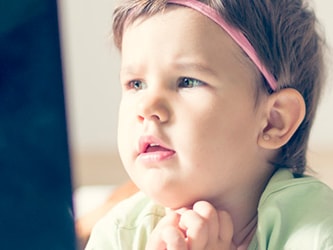 Toddler with a pink headband watches a screen, chin on hands.
