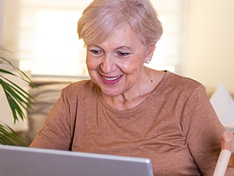 Smiling older woman using a laptop on a couch, surrounded by indoor plants.