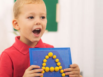 A child holds a blue board with yellow pegs forming "A," watched by an adult, with a plant in the background.