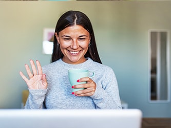 A person smiles, holding a mug and waving at a laptop on a Medbridge video call.