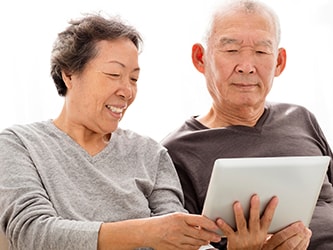 An elderly couple smiles while looking at a tablet on a couch, with a softly lit background.