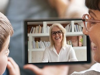 A child and an adult have a video call on a tablet with a smiling woman in glasses in front of a bookshelf, linked to medbridge.