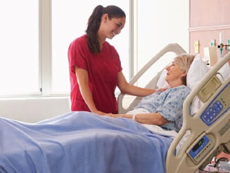 A Medbridge nurse in red scrubs talks to an elderly woman in a hospital bed covered with a blue blanket.