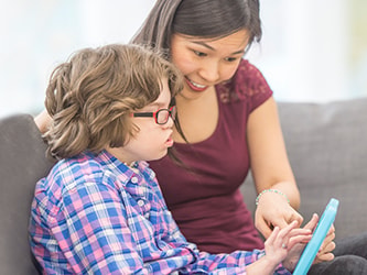 A woman points at a tablet while sitting on a couch with a child in glasses and a plaid shirt.