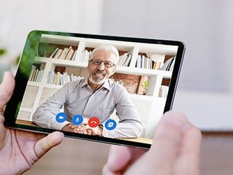 A tablet displays a video call with an older man in glasses, seated by a bookshelf, using medbridge.