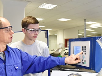 In a brightly lit industrial setting, three people in safety glasses watch a man in blue demonstrate machinery operation at a control panel.