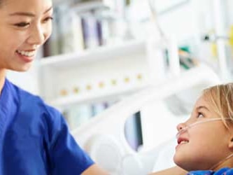 Nurse in blue scrubs smiles while assisting a young girl with an oxygen tube in a hospital bed.