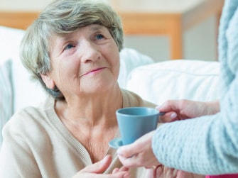 An elderly woman on a sofa is handed a cup of tea by someone standing.