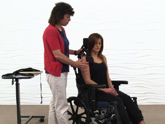 A woman helps adjust another woman's posture in a wheelchair near a table with electronic equipment, promoting Medbridge care.