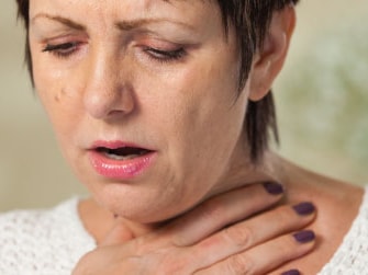 A worried person in a white sweater holds their throat against a blurry green and white background for medbridge.